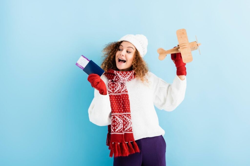 Excited young woman in hat and sweater holding passport and toy plane on blue