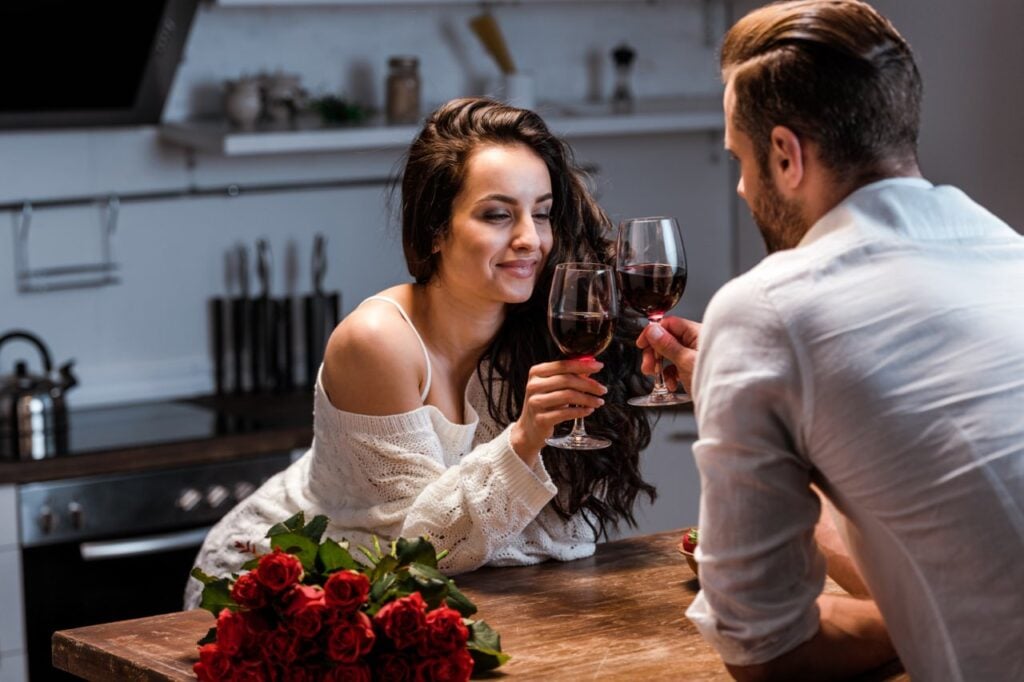 Man and woman clinking with glasses of red wine at wooden table with bouquet of roses