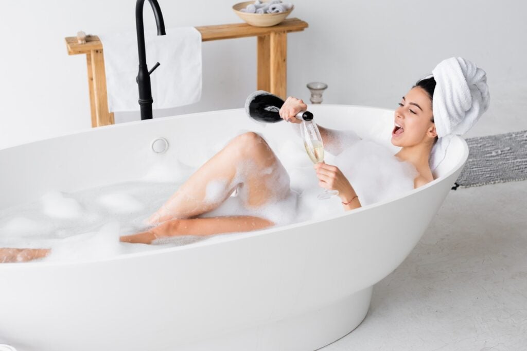 Positive woman pouring champagne in glass while taking bath